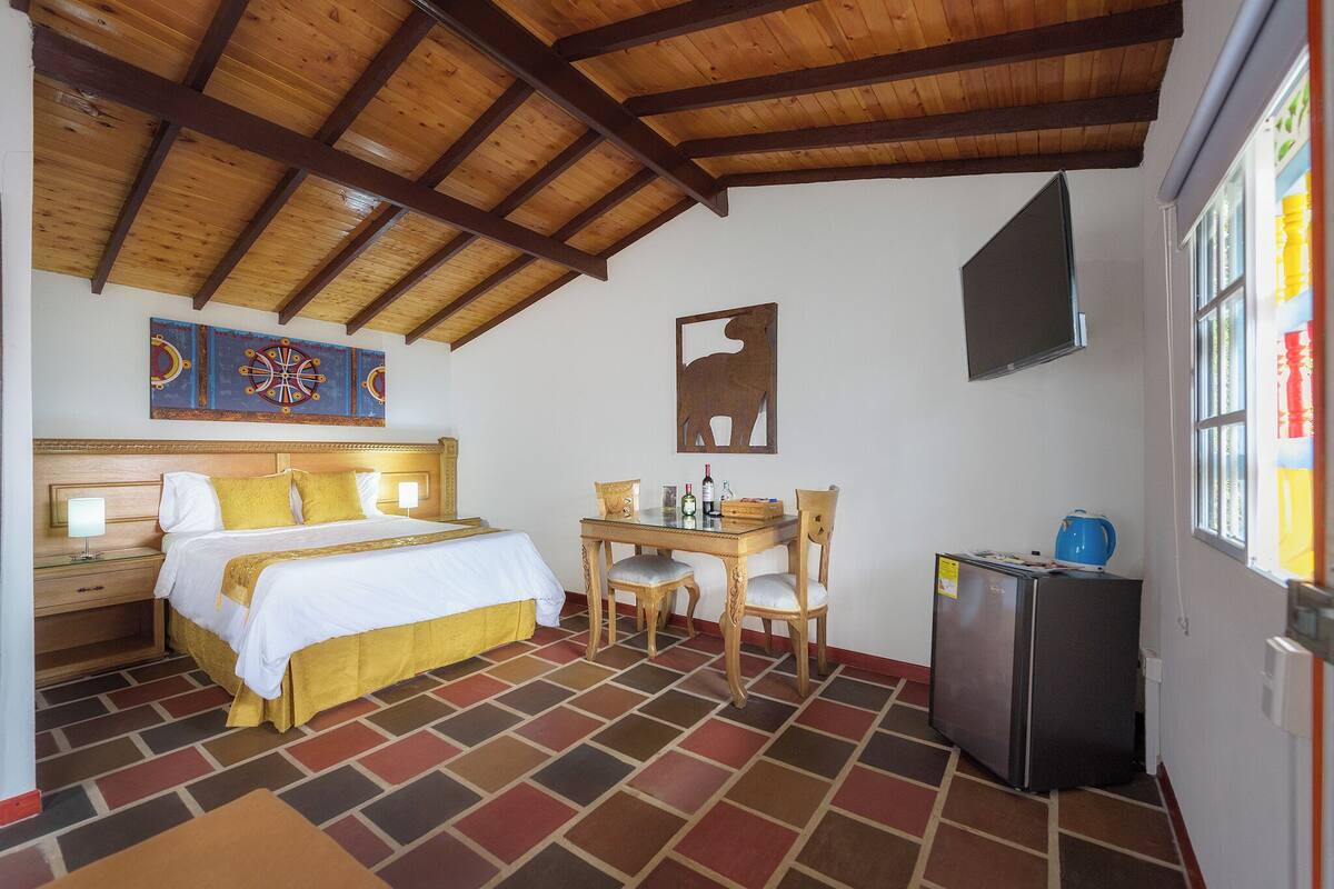 Une chambre d’hôtel avec un plafond en bois, un lit avec une literie blanche, une petite table avec des chaises, un réfrigérateur et un téléviseur fixé au mur.