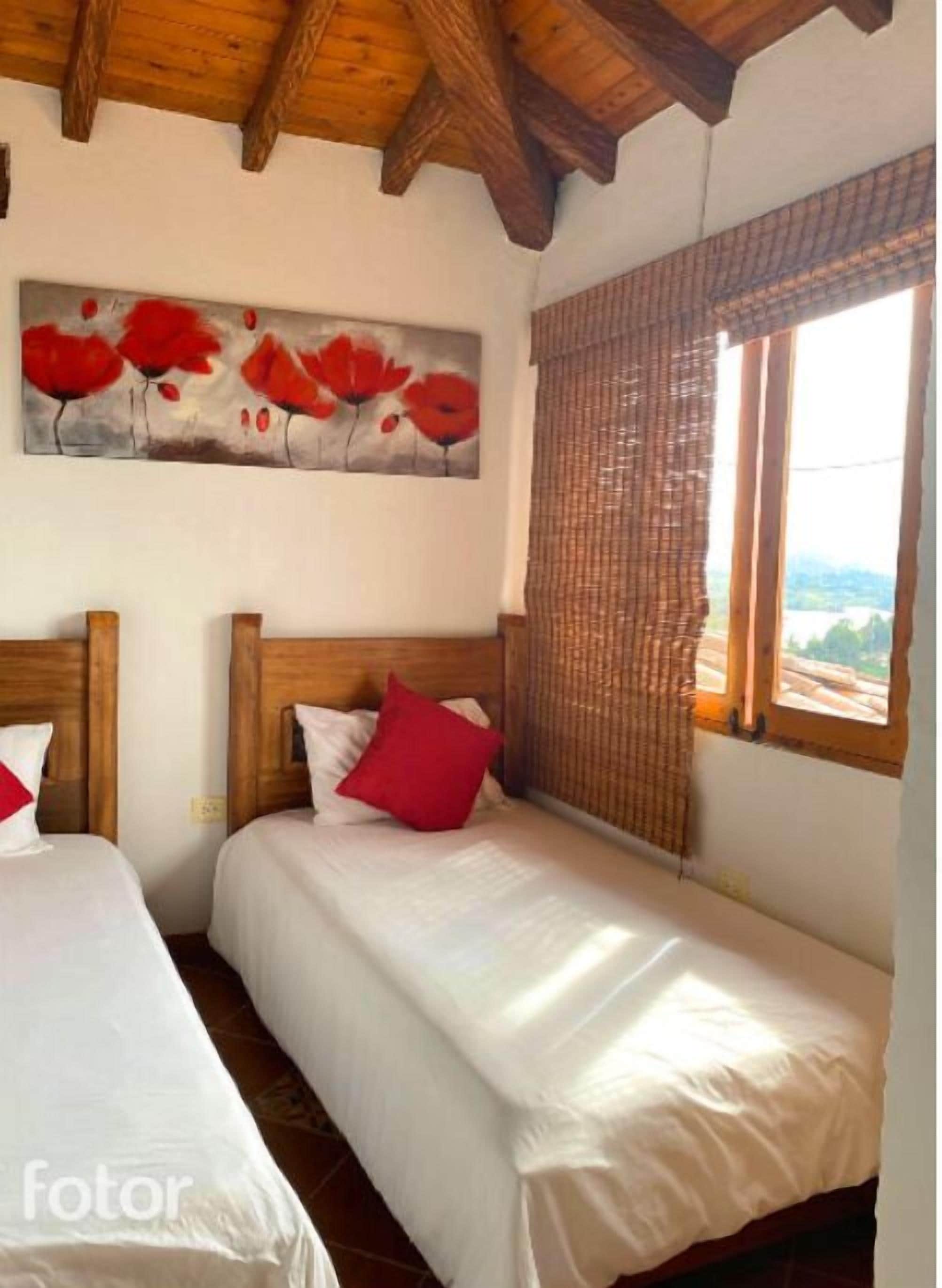 Una habitación con dos camas, cabeceros de madera, un cuadro de flores rojas, una ventana con vistas y una cortina de tejido.