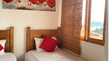 Una habitación con dos camas, cabeceros de madera, un cuadro de flores rojas, una ventana con vistas y una cortina de tejido.