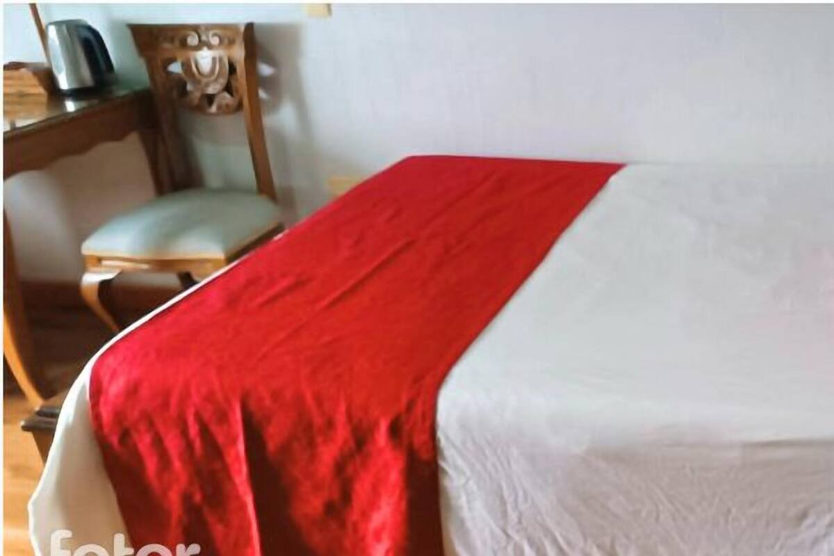 Un lit avec une couverture rouge et des draps blancs, une tête de lit en bois et une chaise avec un coussin.