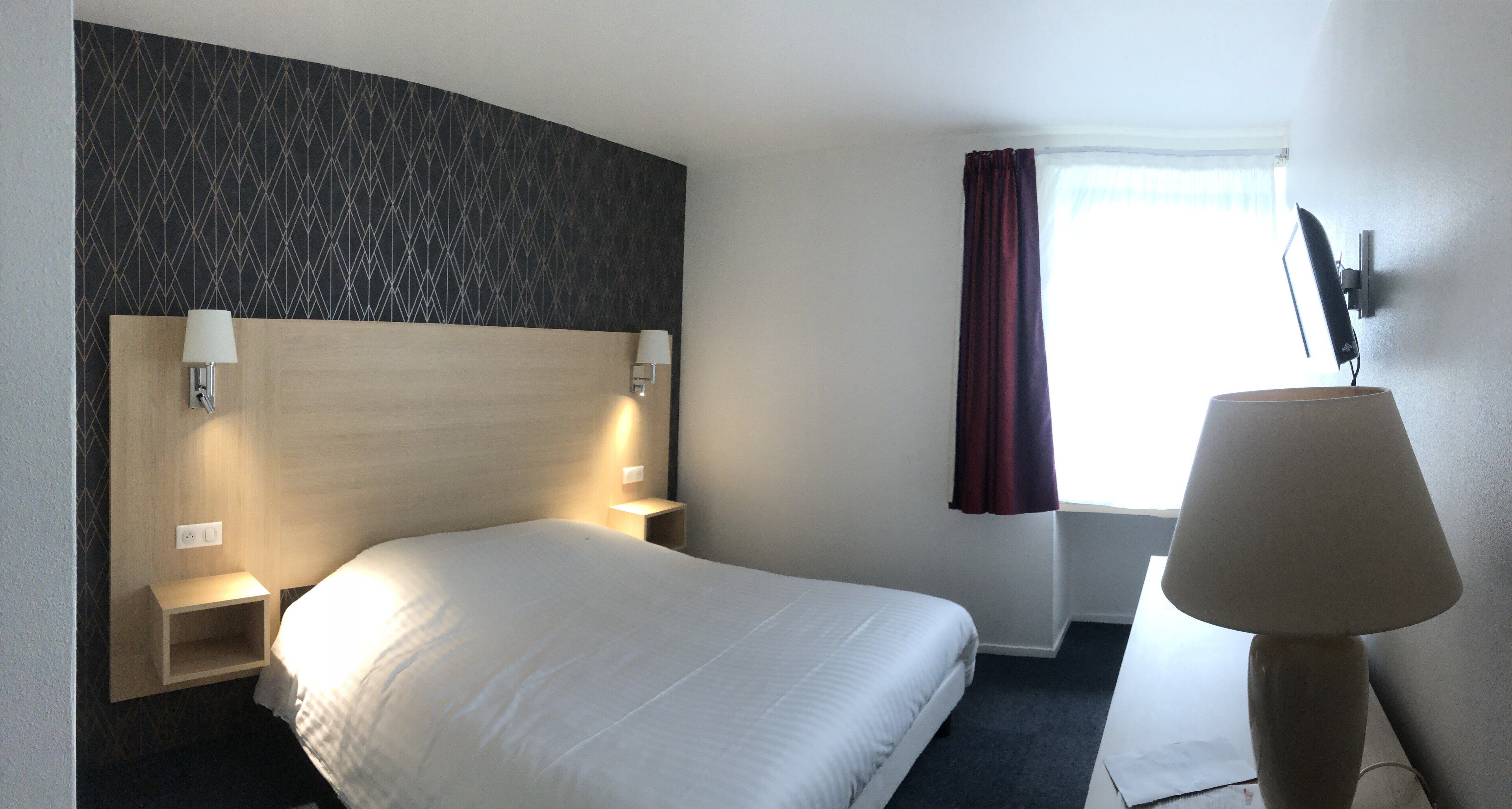 Habitación de hotel con una cama grande, cabecera con diseño geométrico, lámparas de noche, escritorio, silla, ventana con cortinas y televisión montada en la pared.