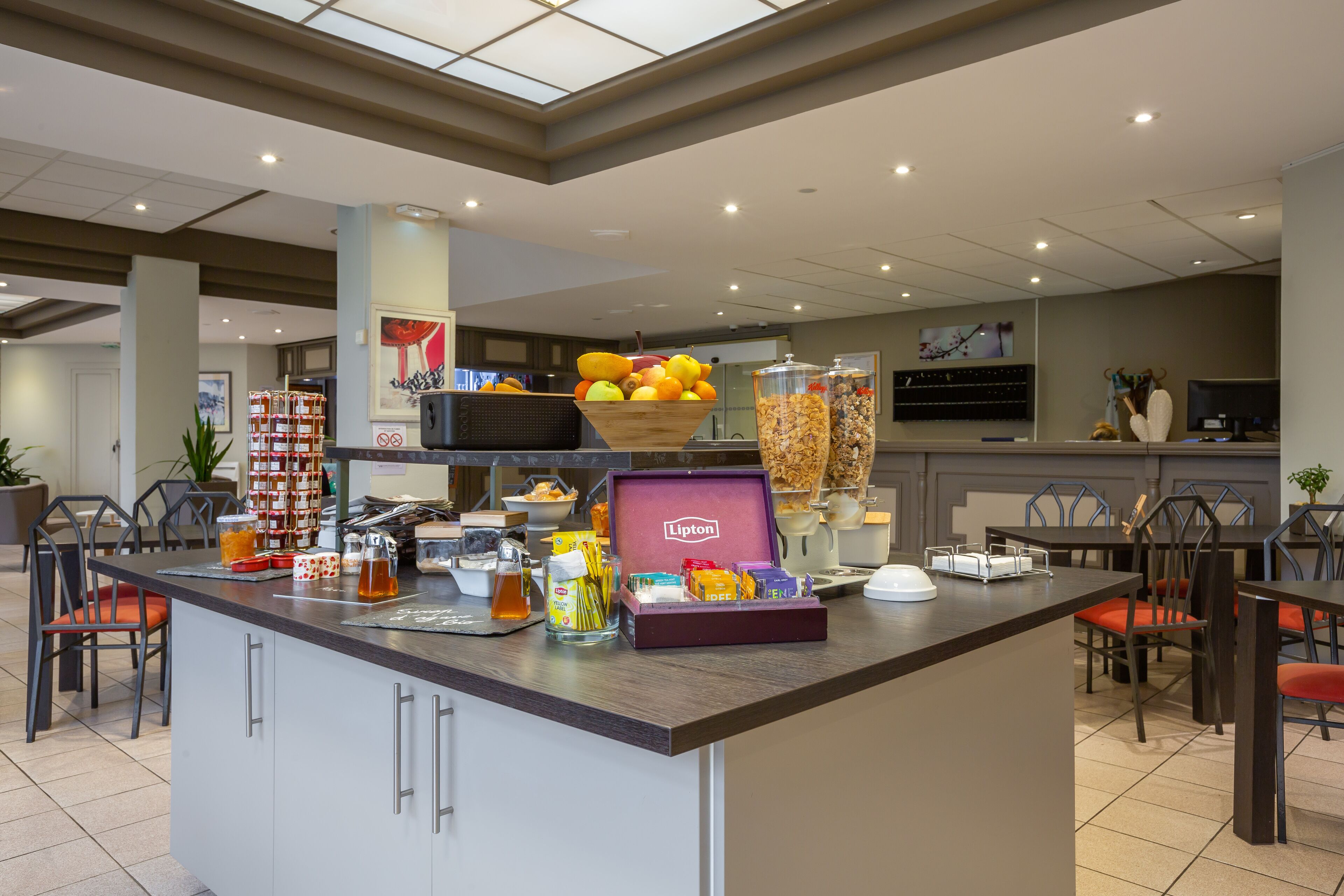 Un espace dédié au petit-déjeuner dans l’hôtel, avec un îlot central où l’on trouve un saladier rempli de fruits, un distributeur de thé Lipton et divers produits alimentaires.