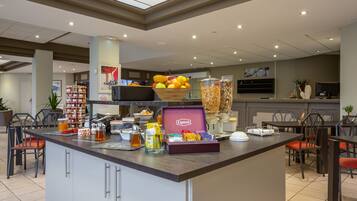 Un espace dédié au petit-déjeuner dans l’hôtel, avec un îlot central où l’on trouve un saladier rempli de fruits, un distributeur de thé Lipton et divers produits alimentaires.
