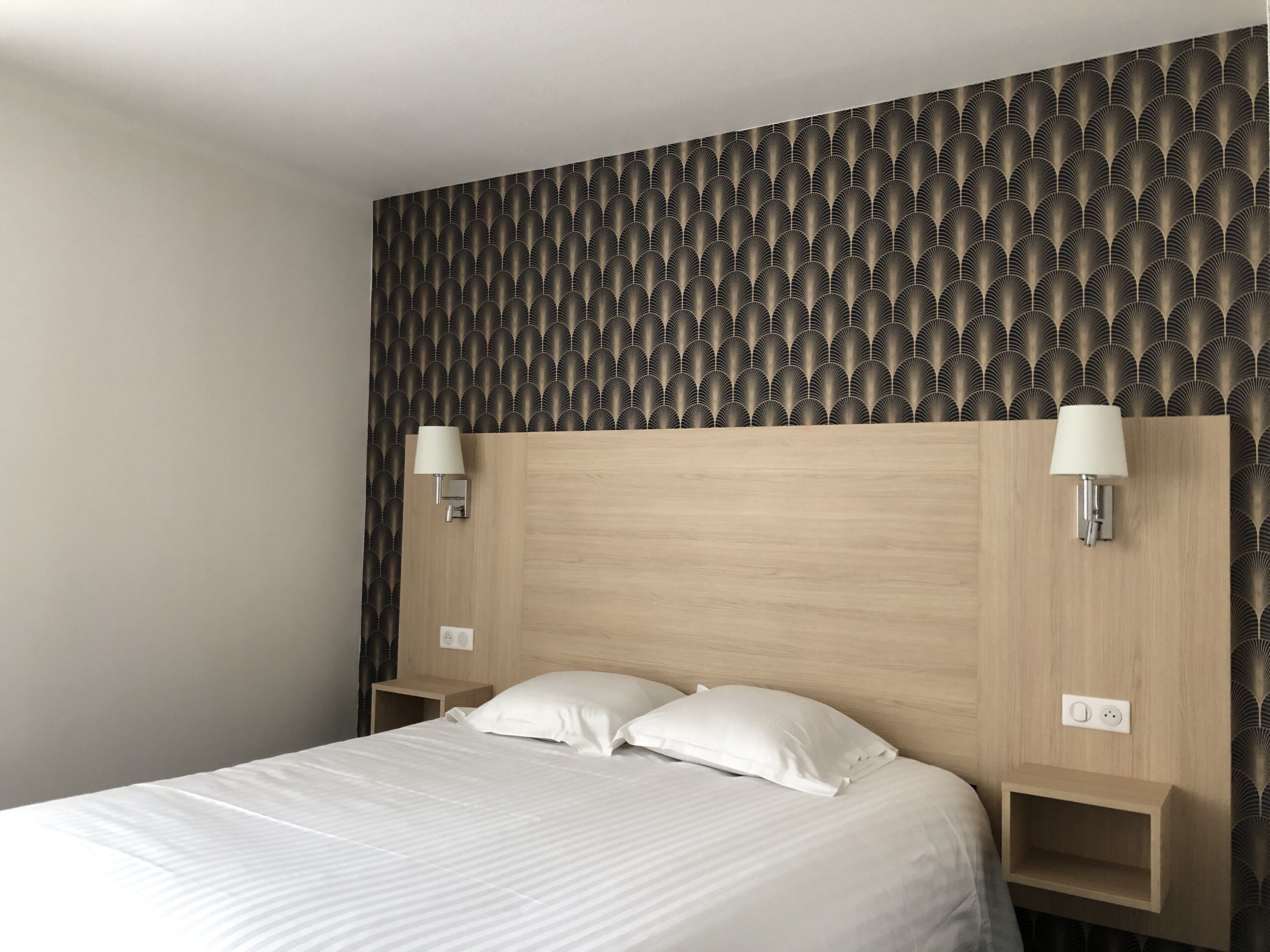 Una habitación de hotel con una cama, dos lámparas de noche, un cabecero de madera y un papel tapiz estampado.