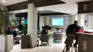 A modern indoor space with a tiled floor, seating areas, and a large screen displaying a sports event. - Cit' Hôtel la Pyramide (Romorantin-Lanthenay)