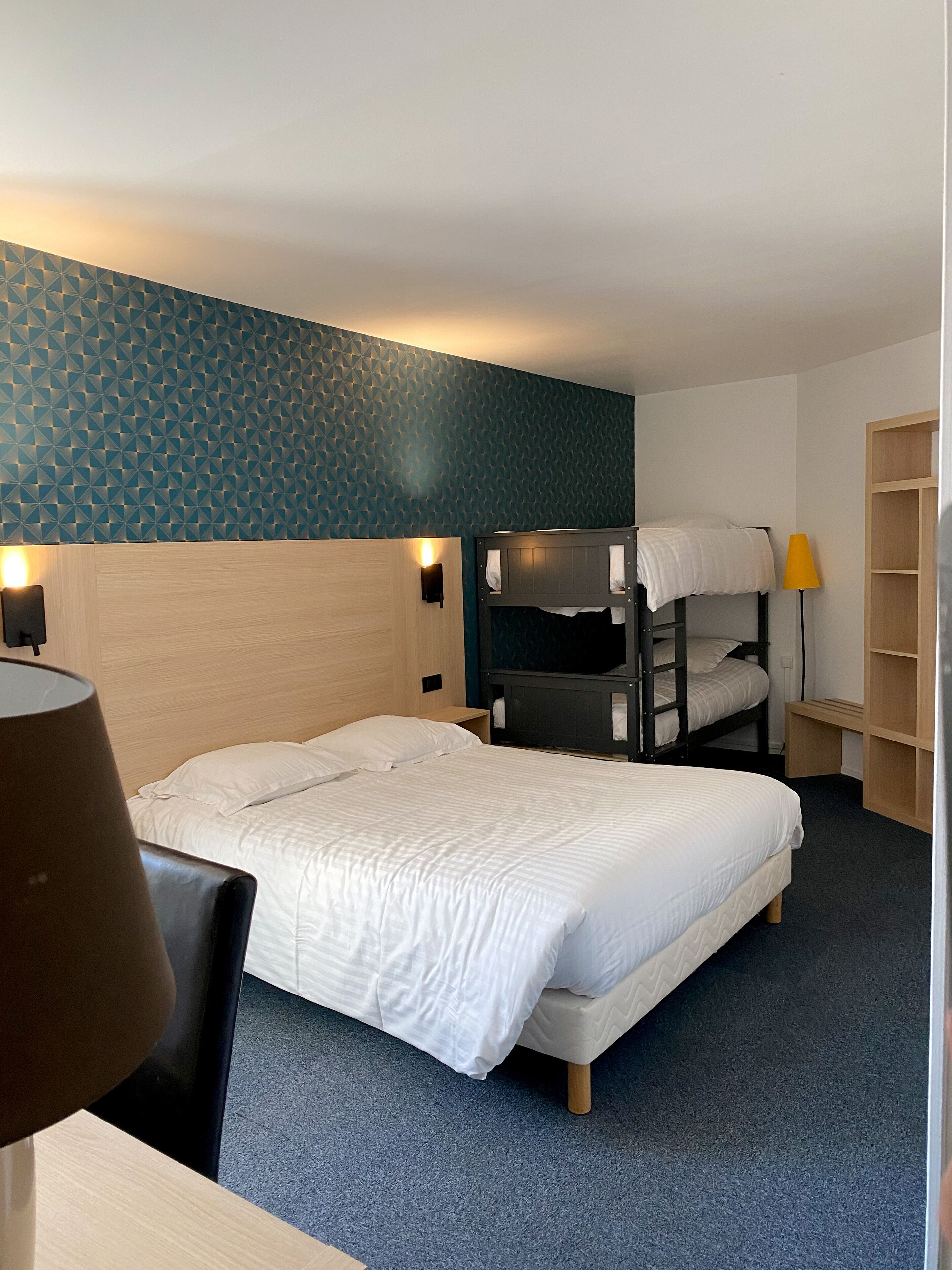 A hotel room with a bunk bed, a single bed, a desk with a lamp, and a bookshelf.