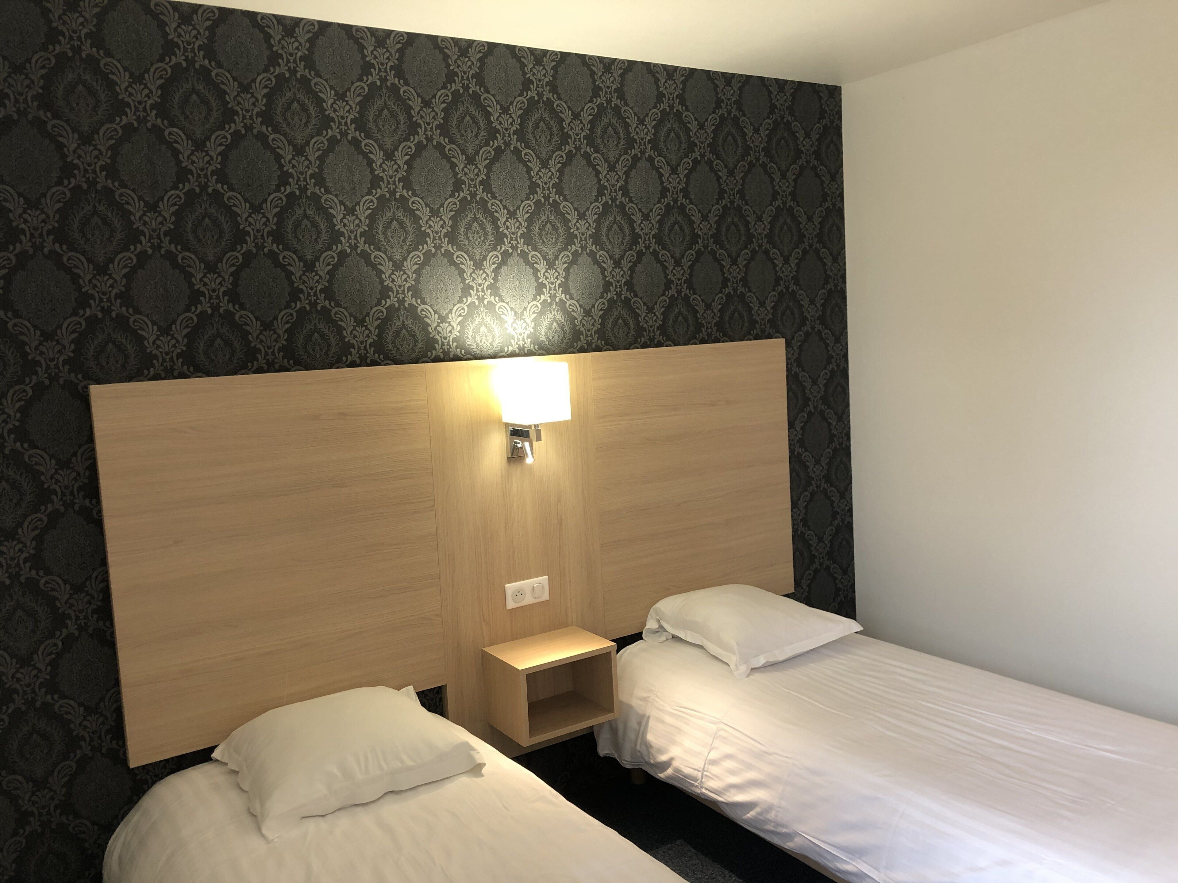 A hotel room with two single beds, a wooden headboard, a nightstand, and a wall-mounted light.