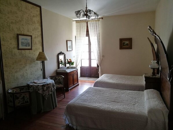 Una habitación con dos camas, un escritorio, una silla, una lámpara y un ventanal con cortinas.