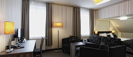 Una habitación de hotel con escritorio, dos sillones, un sofá, una lámpara de pie y una cama con cabecera.