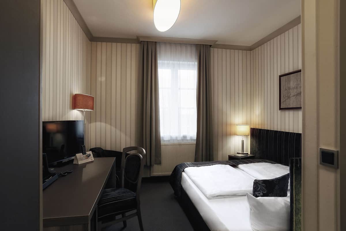Une chambre d’hôtel avec un lit, un bureau équipé d’un ordinateur, une chaise, une télévision et une fenêtre avec des rideaux.