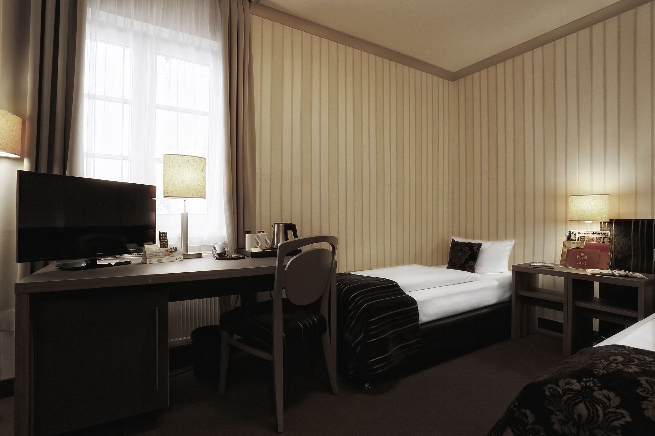 A hotel room with a bed, desk, chair, TV, lamps, and a nightstand with a book and a remote control.