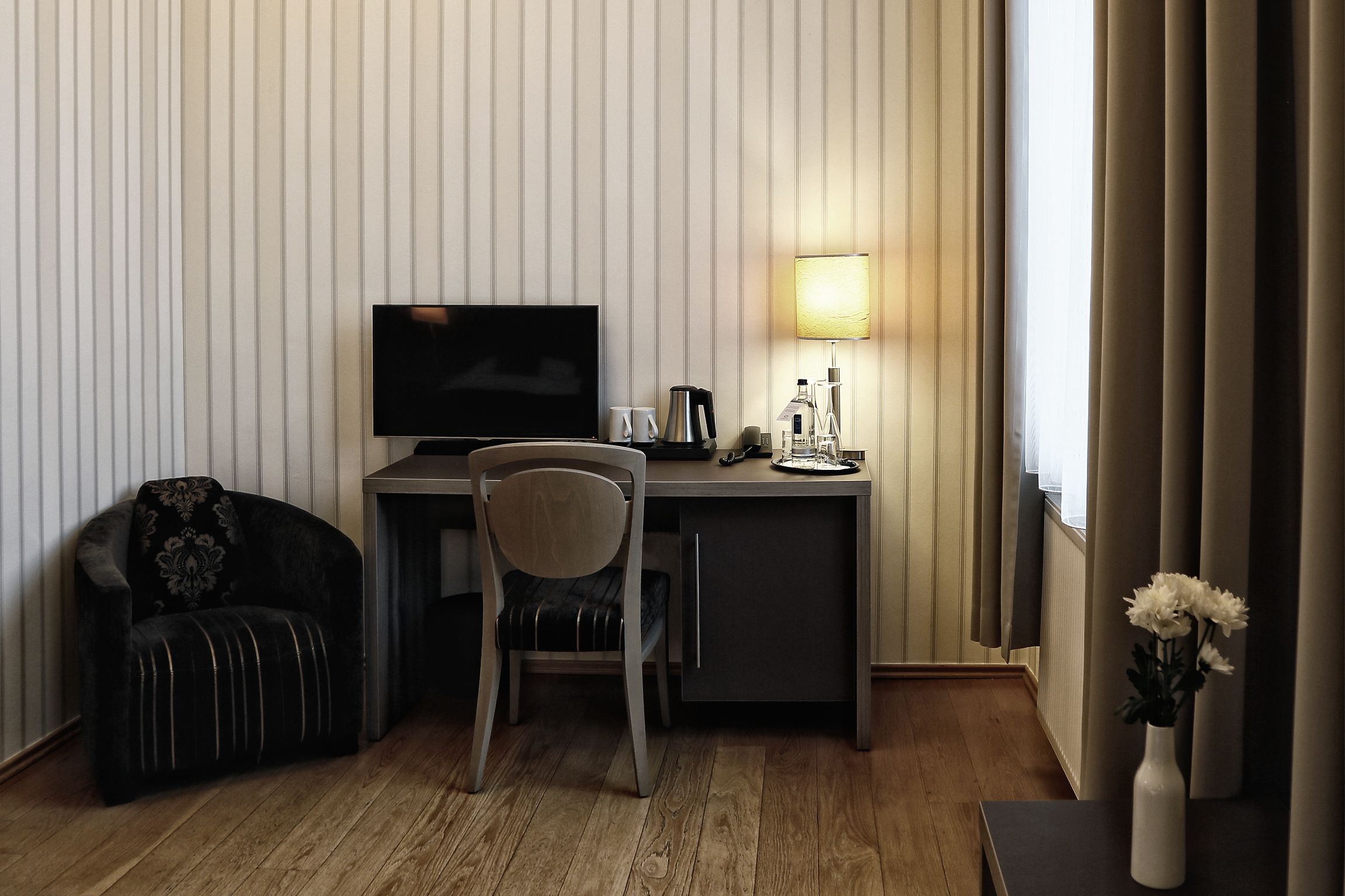 Habitación de hotel con televisión, escritorio, silla y una pequeña mesa con lámpara y flores.