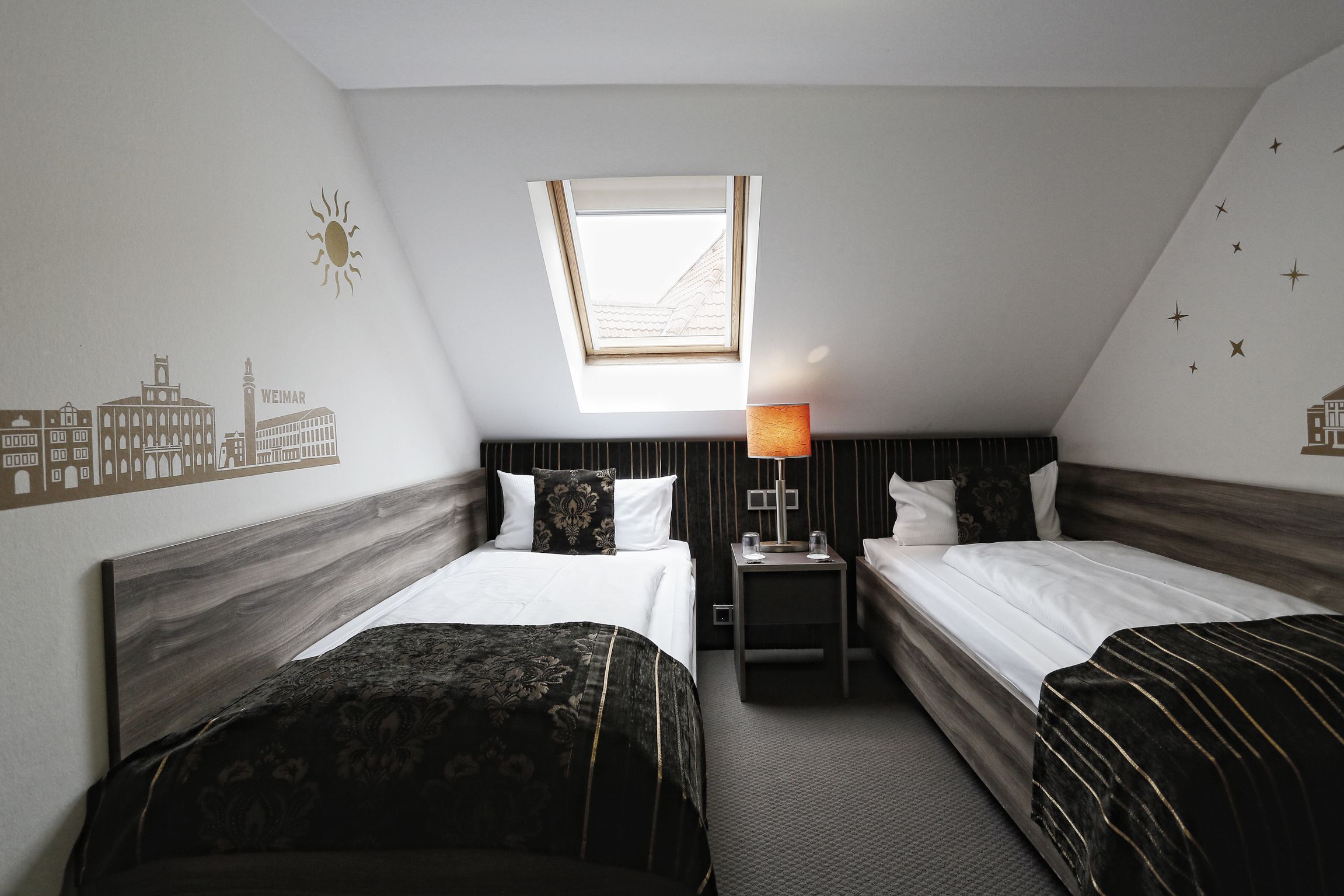 Habitación de hotel con dos camas, tragaluz, lámpara y una vista urbana en la pared.