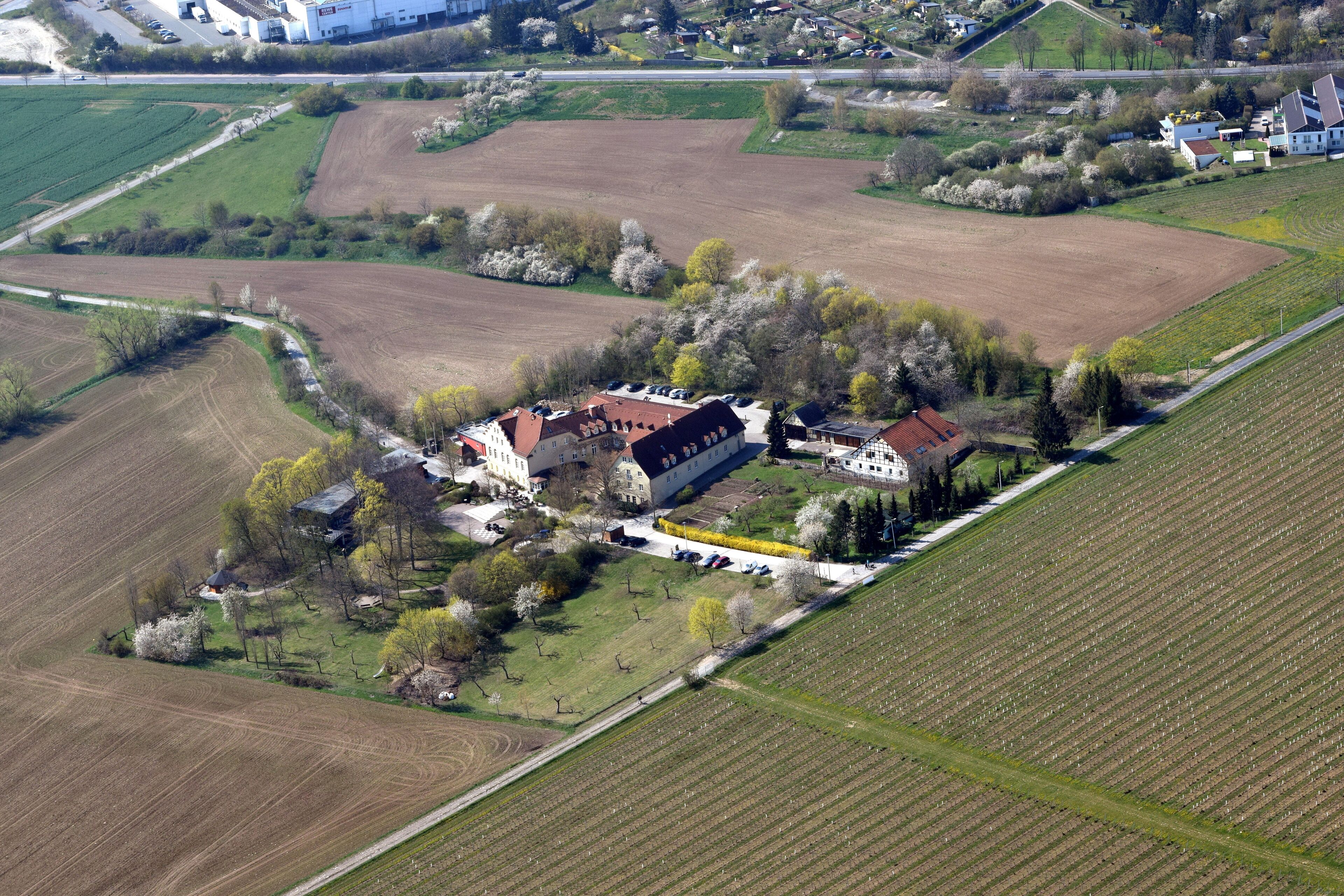 Eine Vogelperspektive auf einen großen Landsitz mit weitläufigen Feldern, einem Hauptgebäude mit rotem Dach und mehreren kleineren Gebäuden.