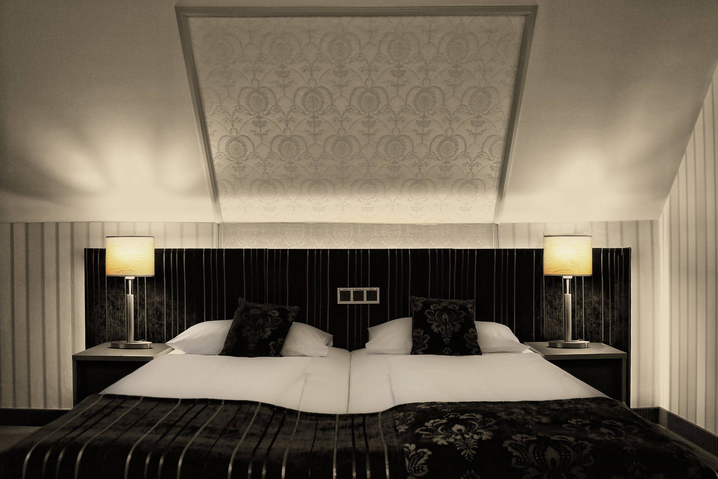 Un grand lit avec une tête de lit sombre, deux tables de chevet avec des lampes et un plafond décoré de motifs.