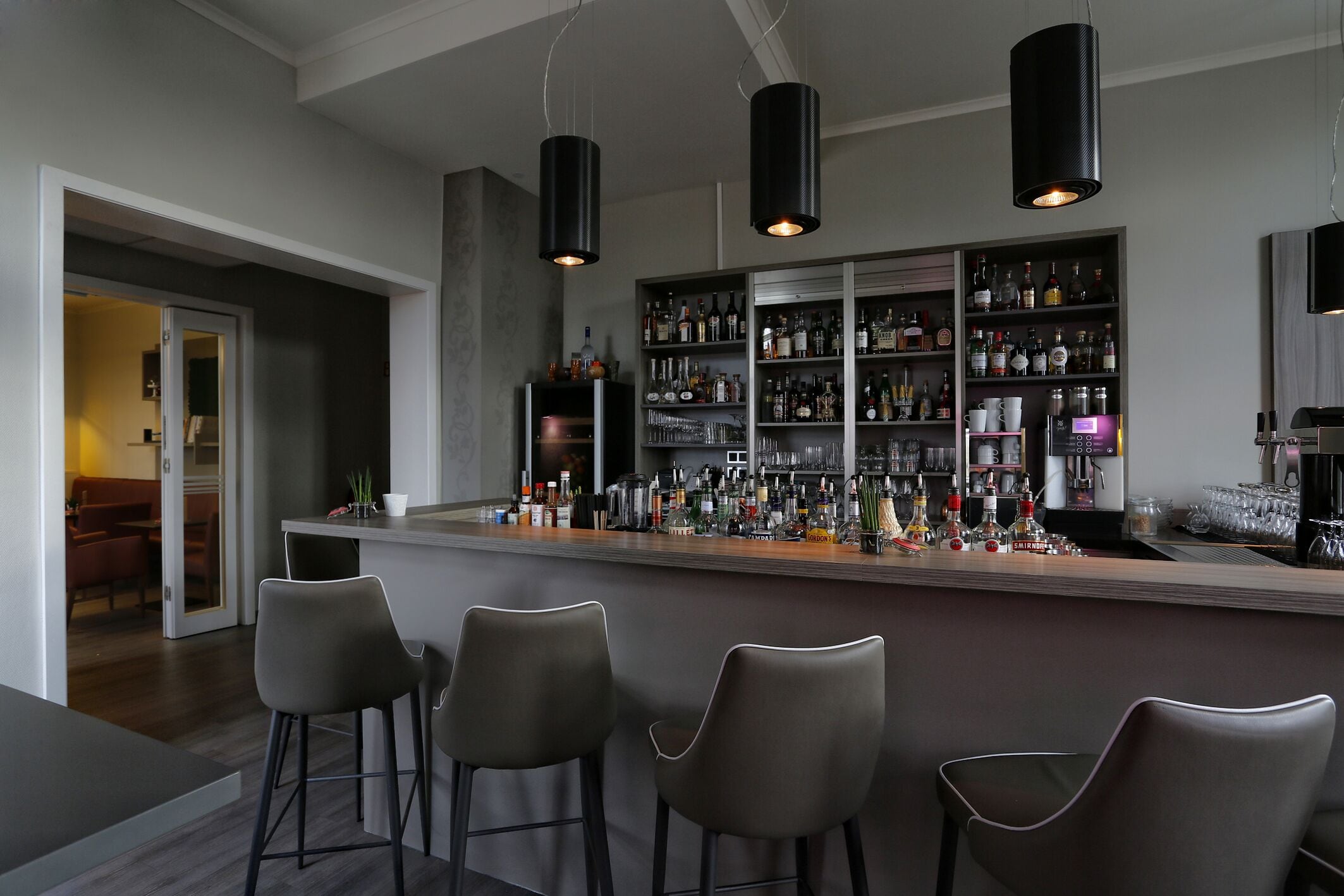 A modern bar area with a bar counter, three stools, and a well-stocked liquor shelf.