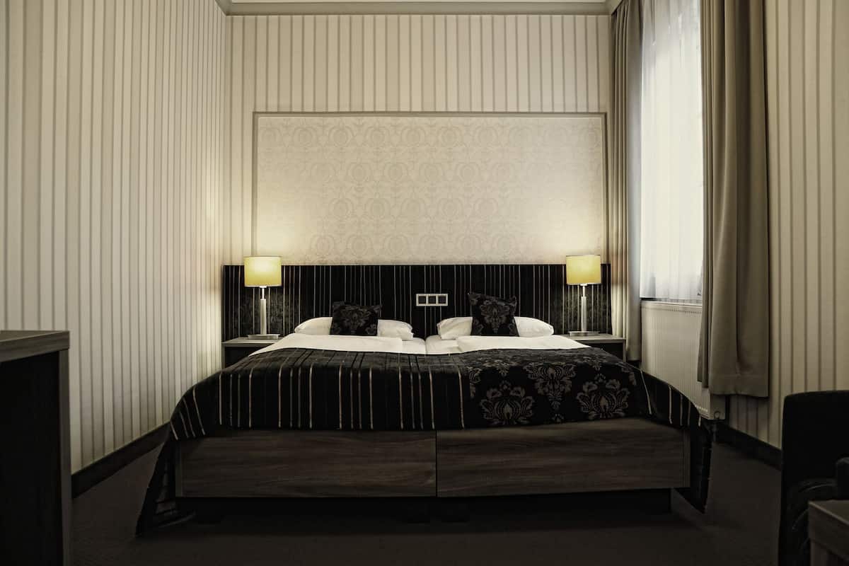 Une chambre d’hôtel avec un grand lit, deux tables de chevet avec des lampes, une tête de lit ornée d’un panneau décoratif et une fenêtre avec des rideaux.