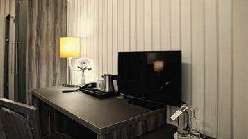 Habitación de hotel con un escritorio de madera, televisor de pantalla plana, lámpara, teléfono, cafetera y minibar con una botella y un vaso.