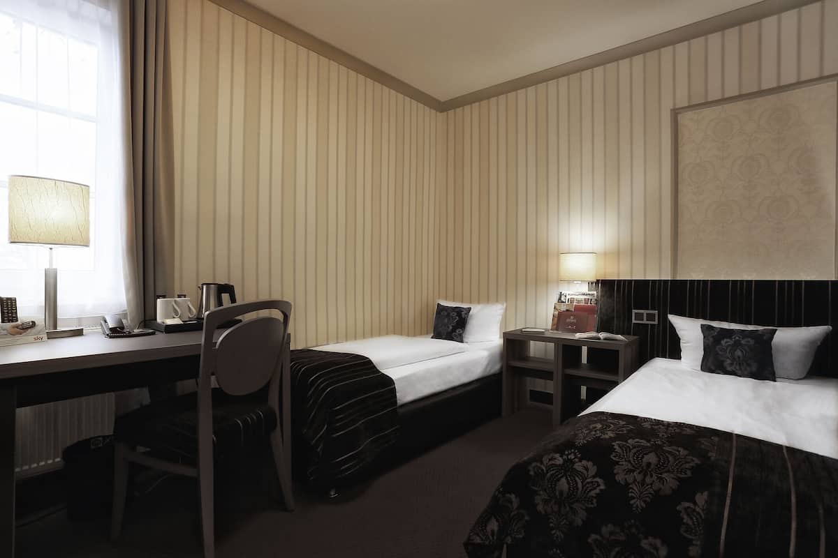 Une chambre d’hôtel avec deux lits, un bureau, une chaise, une lampe, une table de chevet et une fenêtre avec des rideaux.