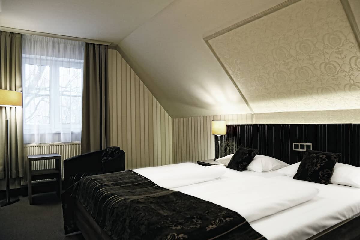 Une chambre d’hôtel avec un grand lit, un bureau et une chaise. Il y a une fenêtre avec des rideaux, un radiateur et une lampe sur le bureau.