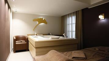 A modern hotel room with a large bed, a small seating area, a bathroom with a large sink and mirror, and a decorative wall piece.