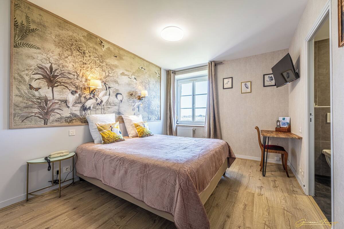 A bedroom with a large bed, a bedside table with a lamp, a wall-mounted TV, and a window with curtains.