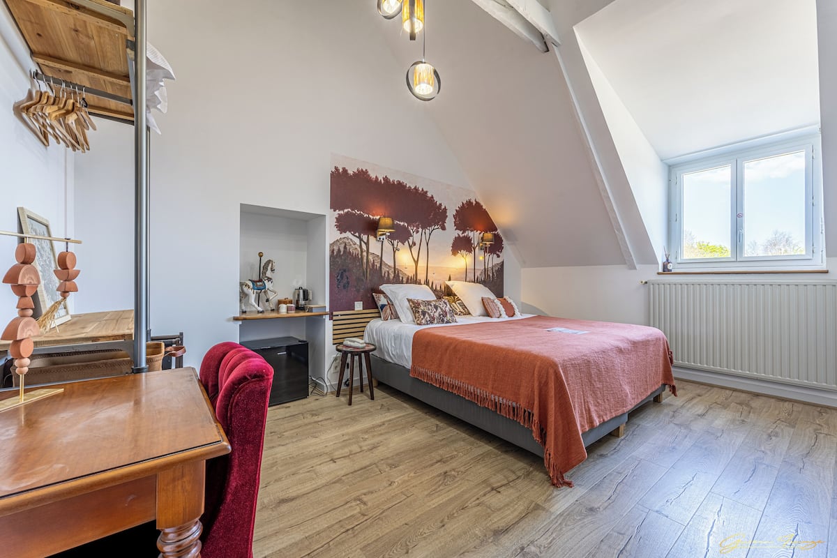 A bedroom with a sloped ceiling, a bed with a red blanket, a desk with a wooden chair, and a window with a view of trees.
