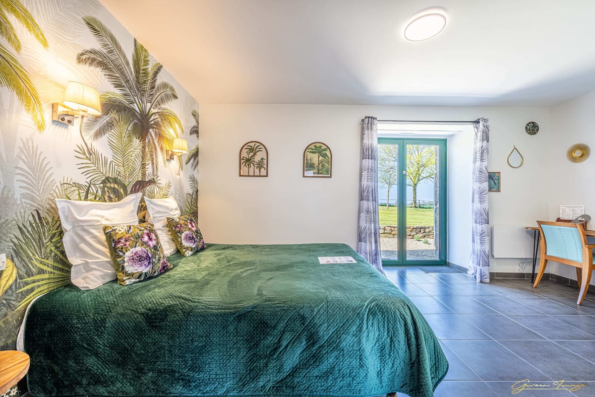 A bedroom with a green bedspread, a wall mural of palm trees, and a view of the outdoors through a glass door.