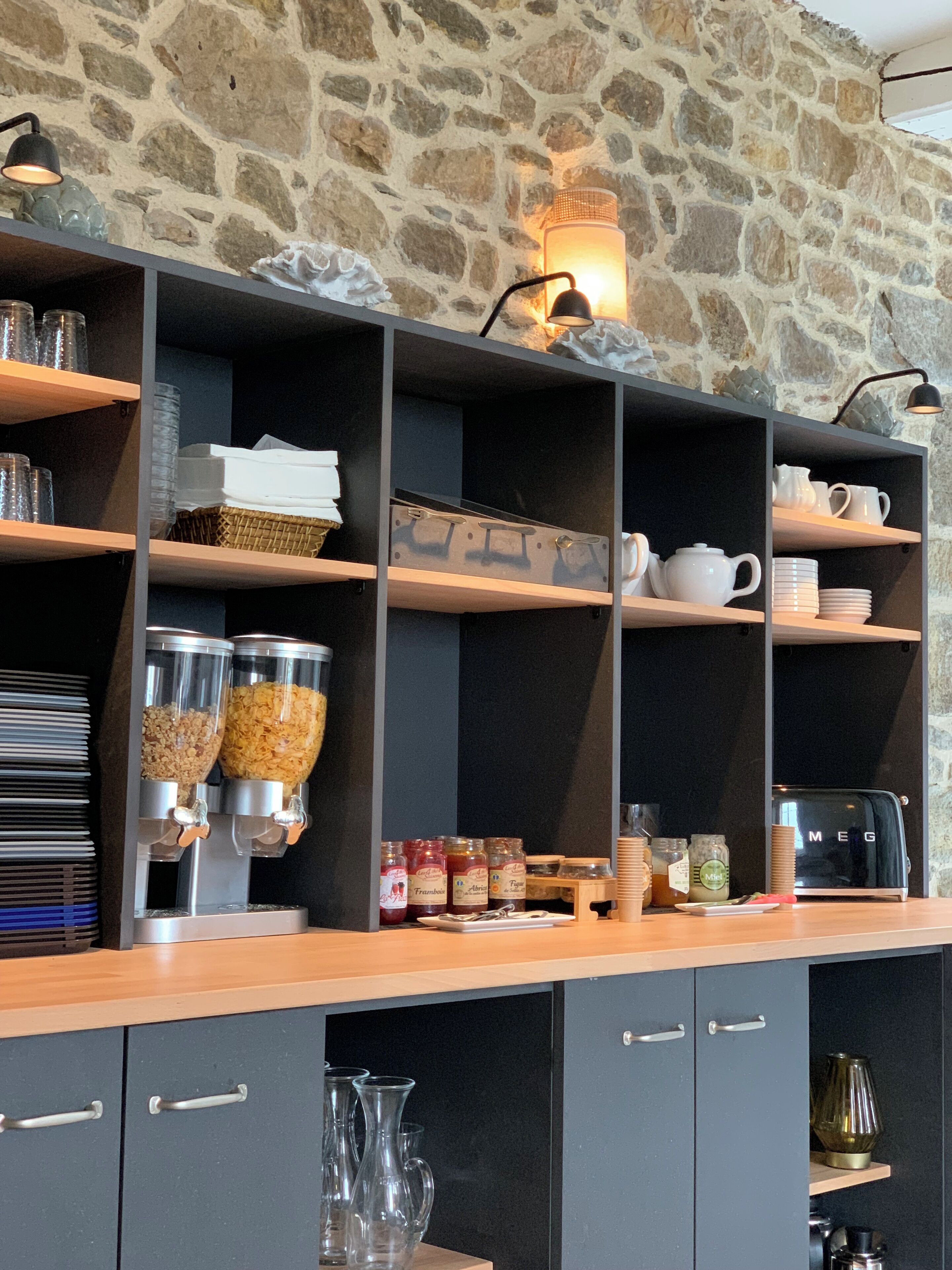 Un bar pour le petit-déjeuner, doté d’étagères où sont rangés divers aliments pour le petit-déjeuner, une machine à café et un mur en pierre en arrière-plan.