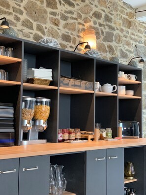 A breakfast bar with shelves holding various breakfast items, a coffee machine, and a stone wall in the background. - Manoir de Rigourdaine (Plouer-sur-Rance)