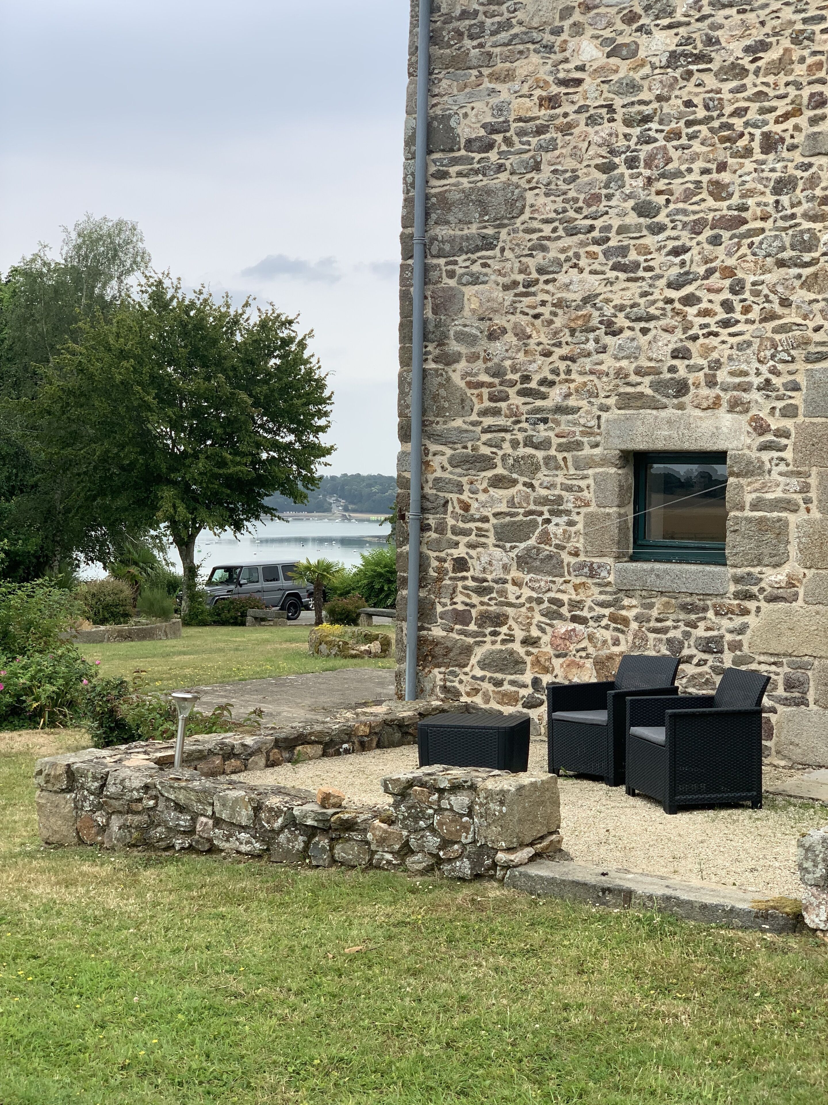 Un bâtiment en pierre offrant une vue sur l’eau, avec des places assises à l’extérieur et un VUS garé à proximité.