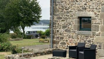Un bâtiment en pierre offrant une vue sur l’eau, avec des places assises à l’extérieur et un VUS garé à proximité.