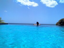 Uma pessoa sentada em uma plataforma flutuante em uma piscina azul translúcida, com vista para o oceano e uma ilha ao longe.