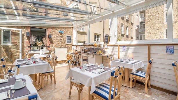 A restaurant with outdoor seating, stone walls, and a glass roof. Tables are set with white tablecloths and blue-striped chairs. - Hôtel Quic en Groigne (Saint-Malo)