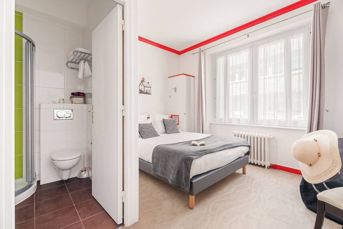 A modern bedroom with a bed, a window with curtains, a radiator, and a bathroom with a toilet and shower.