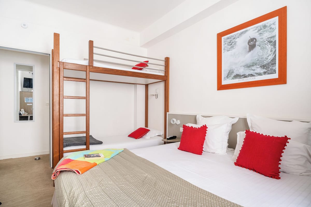 A room with a bunk bed, a desk with a chair, a bed with white and red pillows, and a framed picture on the wall.