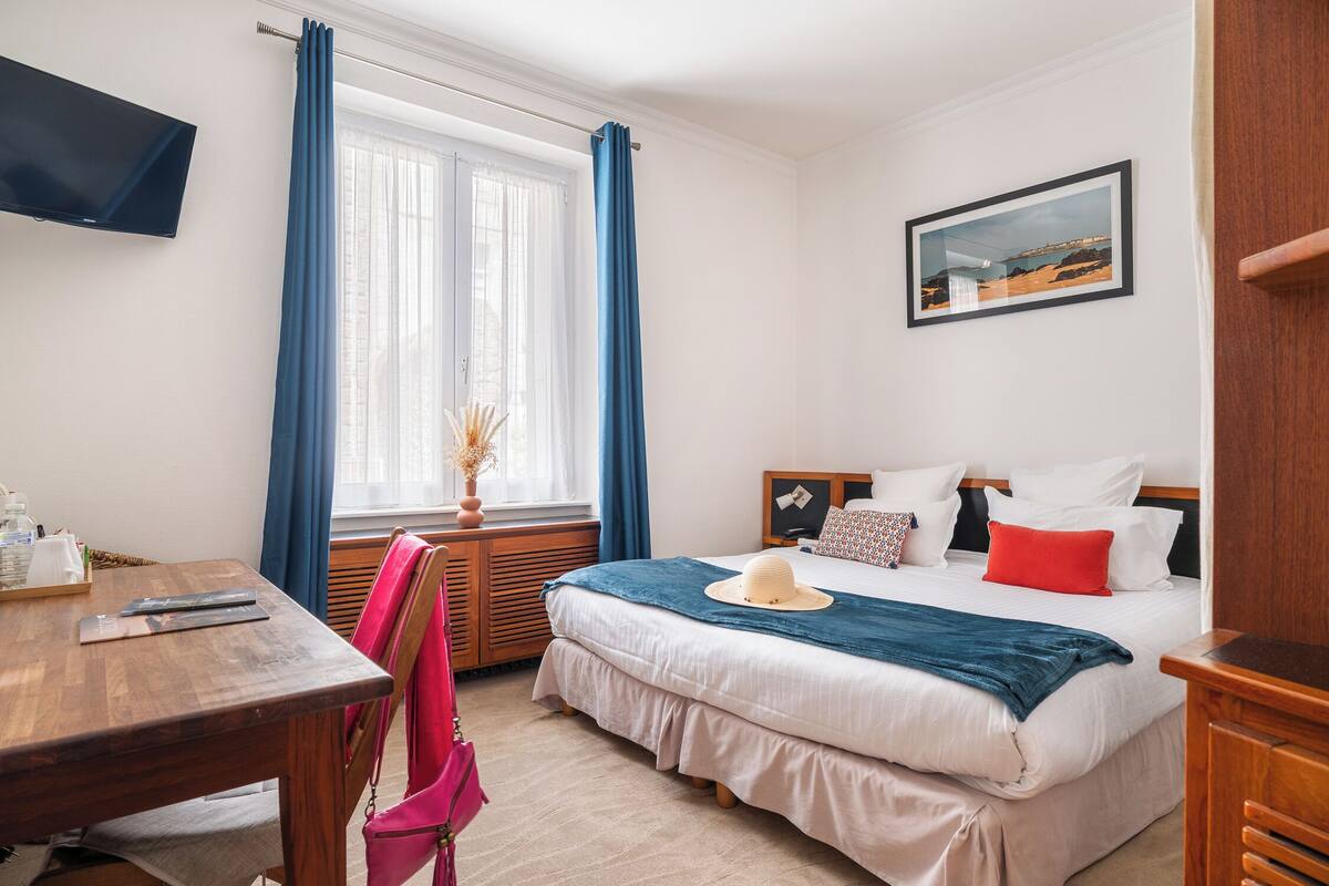 A hotel room with a bed, a desk, a chair, a TV, and a window with curtains.