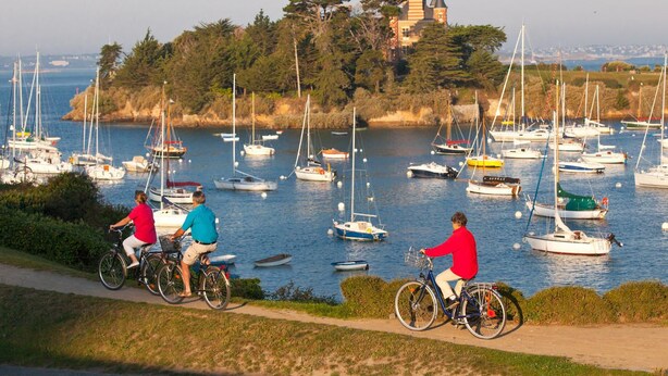 Un paysage côtier avec une marina remplie de voiliers et de yachts, une piste cyclable et un bâtiment historique situé sur une colline en arrière-plan.