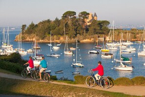 Un paysage côtier avec une marina remplie de voiliers et de yachts, une piste cyclable et un bâtiment historique situé sur une colline en arrière-plan.