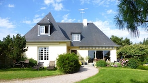 Une maison à deux étages, dotée d’un toit en pente, d’un porche couvert et d’une allée en gravier menant à une pelouse aménagée avec du mobilier d’extérieur.