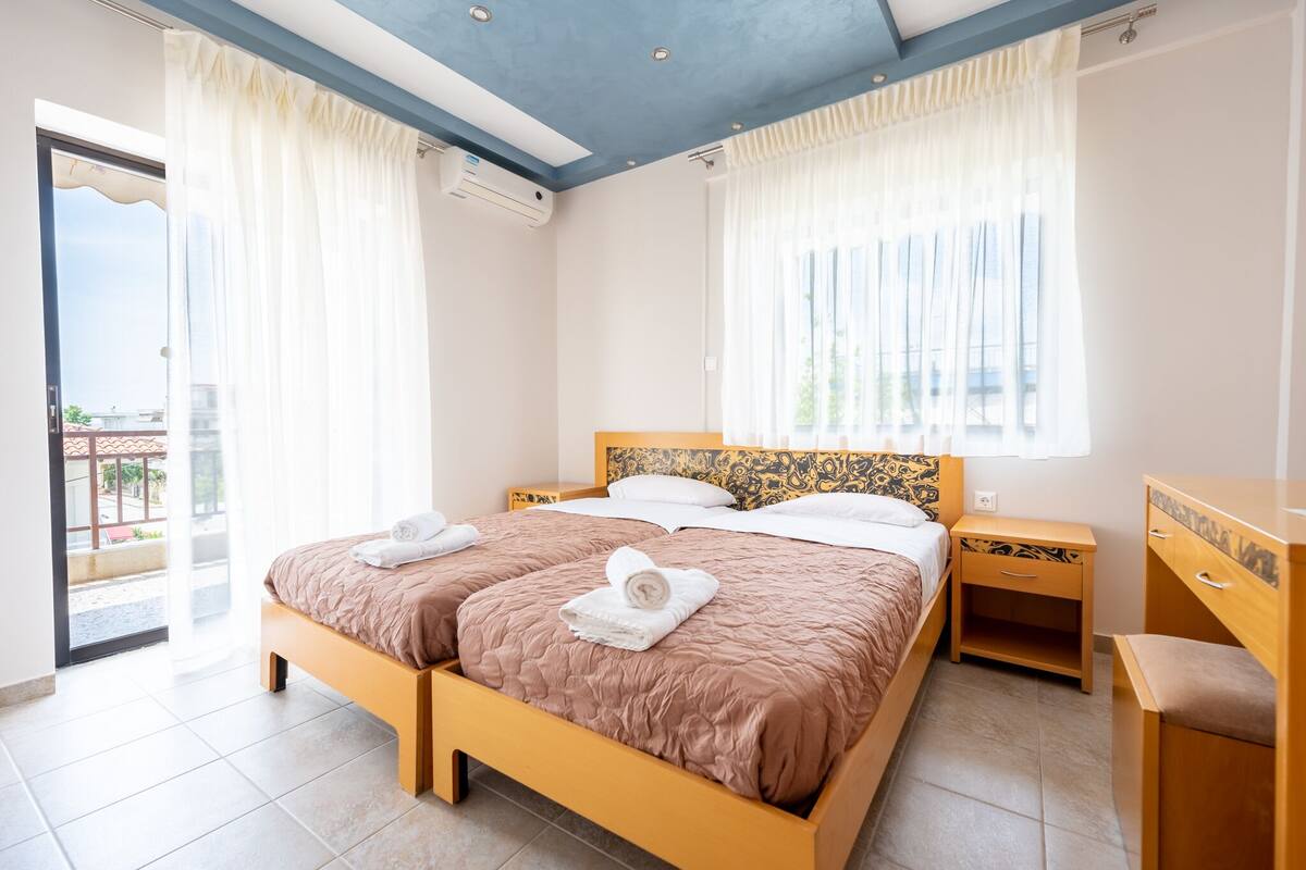 A double bed with a brown quilt, two white towels, a wooden nightstand, a wooden dresser, and a balcony door.