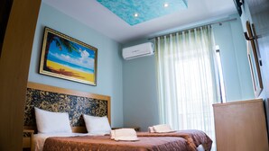 A hotel room with two beds, a ceiling with a skylight, a painting of a beach, an air conditioning unit, and a window with curtains. - Milonas House Apartments (Nea Propontida)