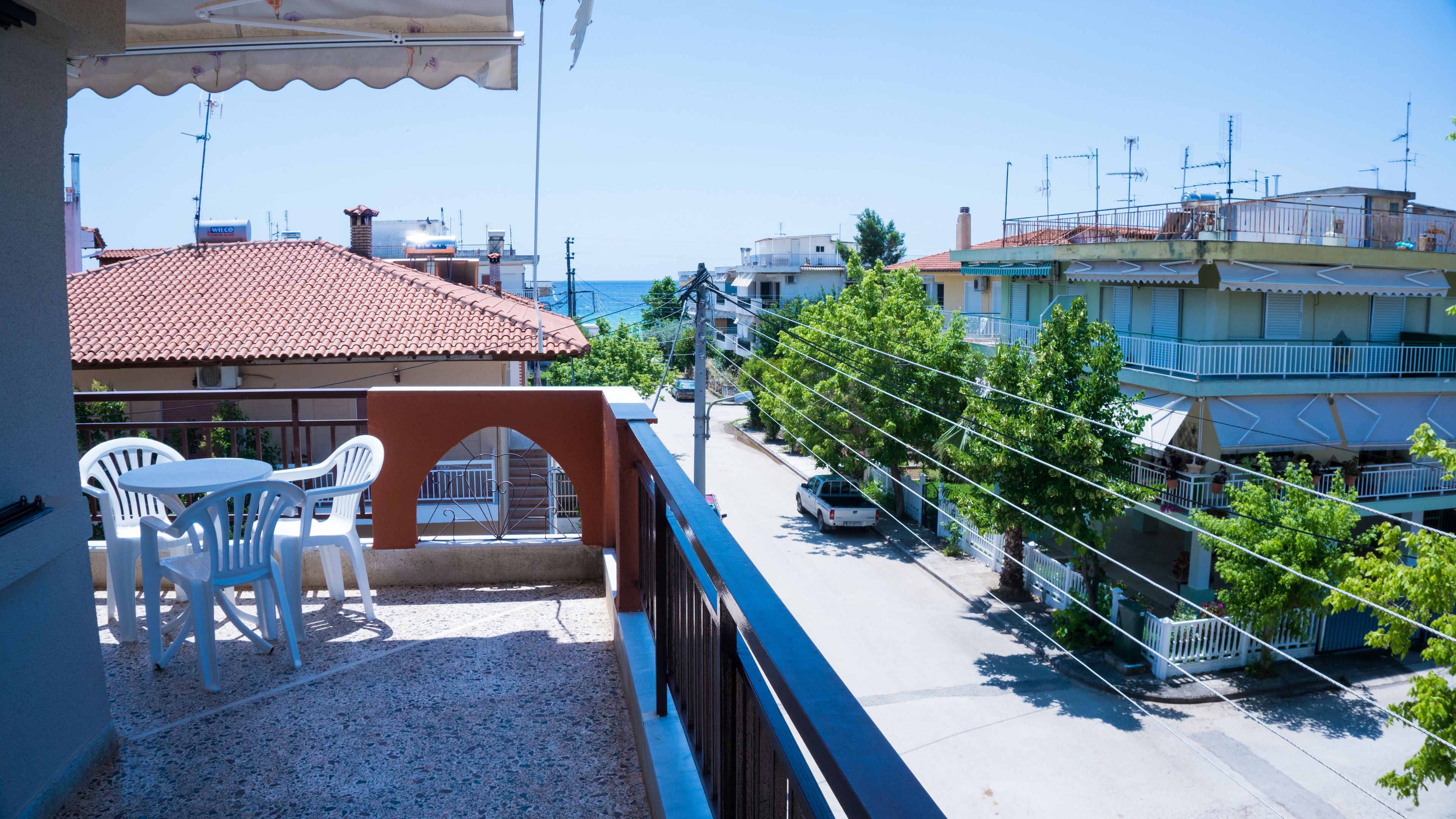 A balcony with white plastic chairs and a table, overlooking a street with buildings and a clear sky.