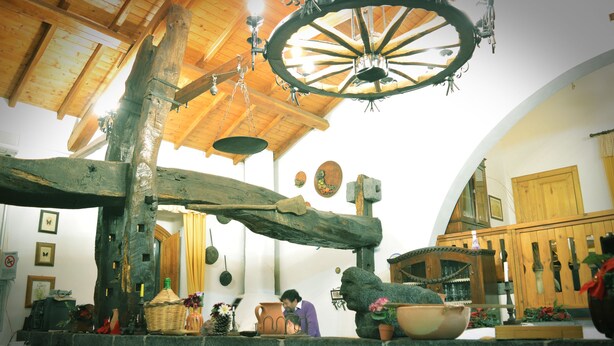 Ein rustikales Interieur mit Holzbalkendecke, einer großen Wagenleuchter-Lichtinstallation und einer Steinwand mit diversen Gegenständen.