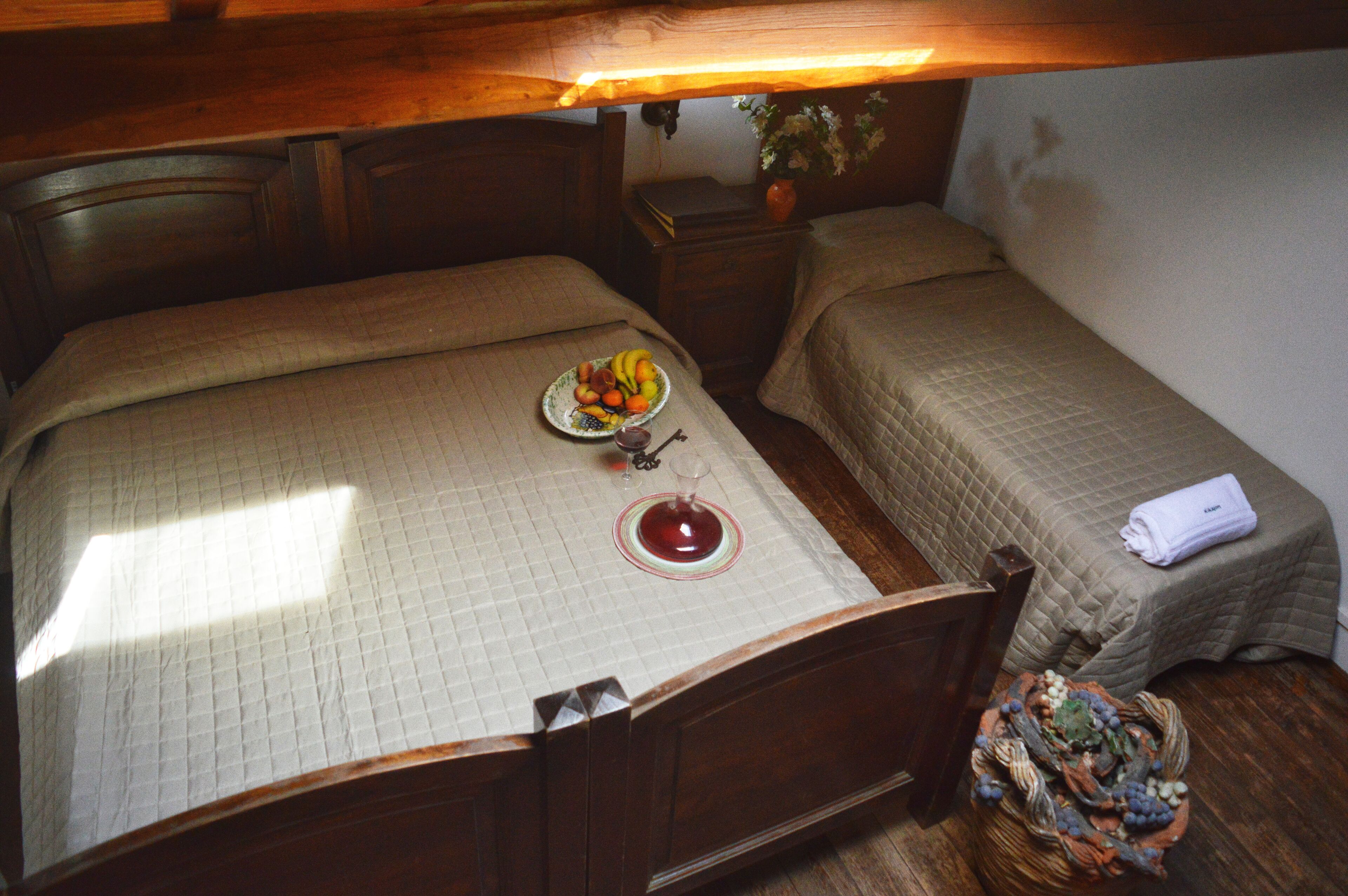 A small, cozy bedroom with two beds, a bedside table with a vase, and a basket of fruit on the floor.