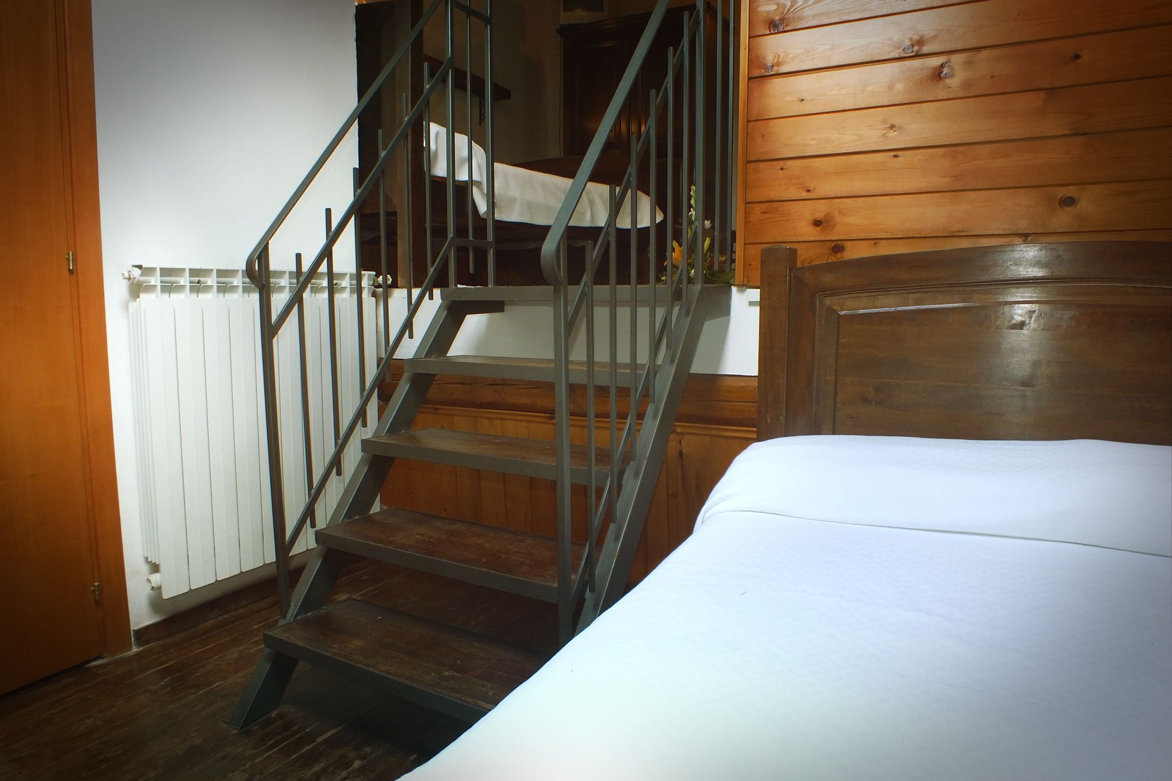 A bedroom with a wooden headboard, a bed, a staircase with metal railings, and a radiator.