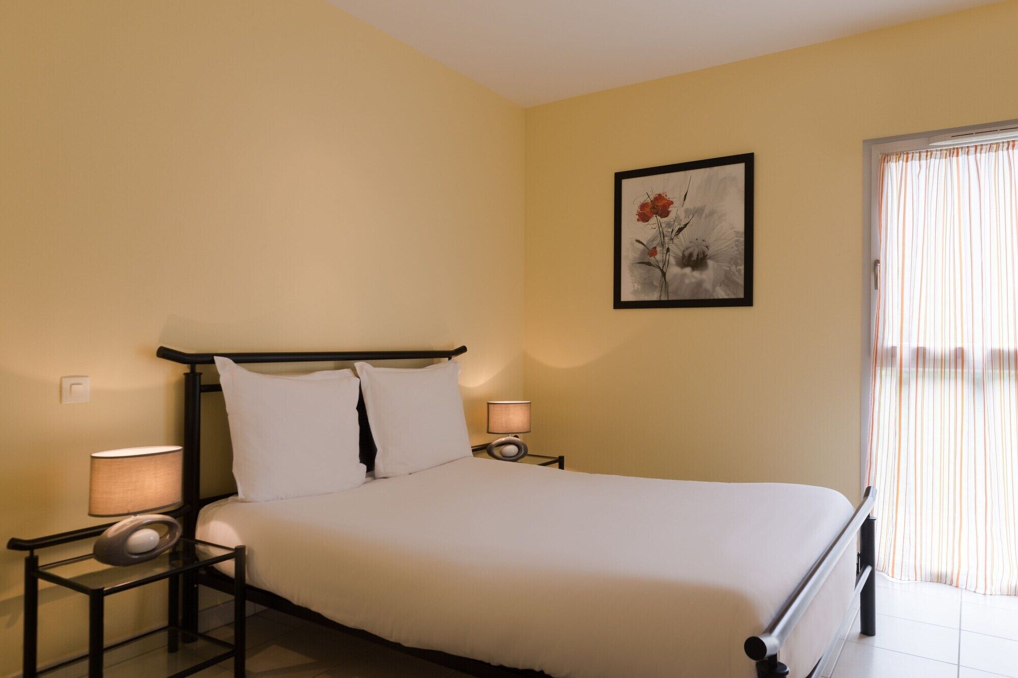 A bedroom with a bed, bedside lamps, a nightstand, a framed artwork on the wall, and a window with curtains.
