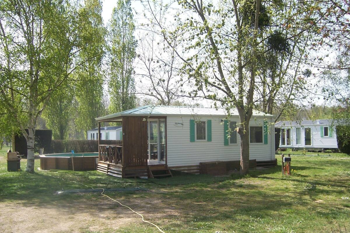 Ein Mobilheim mit Veranda und kleiner Terrasse, umgeben von Bäumen und Gras.