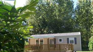 A single-story mobile home with a wooden deck, surrounded by a grassy yard and dense trees. - Camping les Terrasses de Rivarennes (Rivarennes)