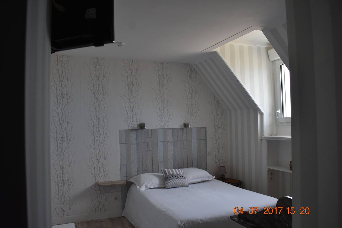 A bedroom with a sloped ceiling, a bed with white bedding, a nightstand, a wall-mounted TV, and a window with blinds.